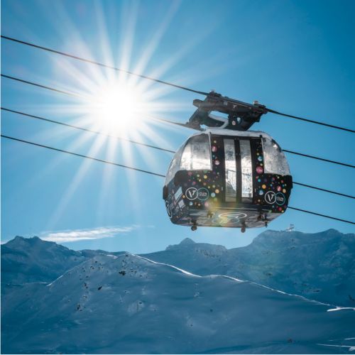 Télécabine décorée suspendue au-dessus d'un paysage alpin enneigé sous un ciel bleu clair avec un soleil éclatant.