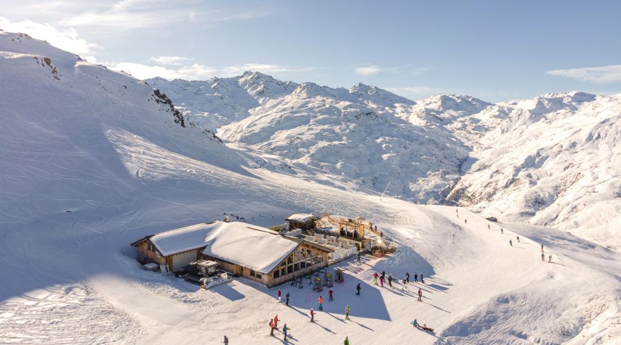 Vue aérienne d'un chalet en bois au sommet enneigé d'une montagne avec des skieurs et snowboardeurs autour, entouré de montagnes enneigées sous un ciel clair.