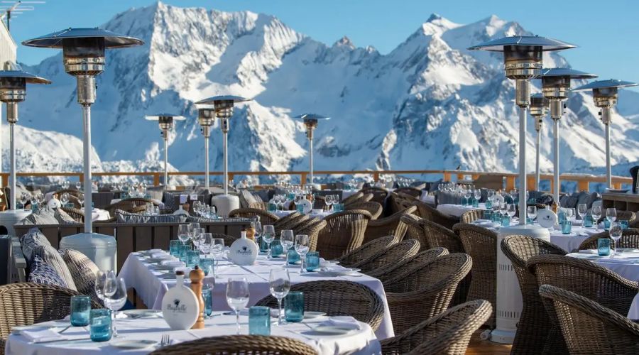 Terrasse d'un restaurant avec des tables dressées de nappes blanches, verres, salières et poivrières, entourées de chaises en osier, avec des chauffages extérieurs et une vue sur des montagnes enneigées sous un ciel bleu clair.