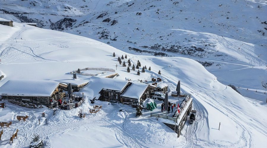 Vue aérienne d'un chalet en bois entouré de neige dans une station de montagne avec quelques personnes sur la terrasse, des sapins dispersés et des pistes de ski visibles dans le paysage enneigé.