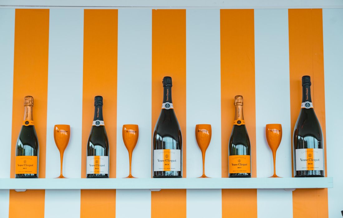 Quatre bouteilles de champagne Veuve Clicquot alternant avec quatre flûtes à champagne orange sur une étagère blanche devant un mur à rayures orange et blanc.