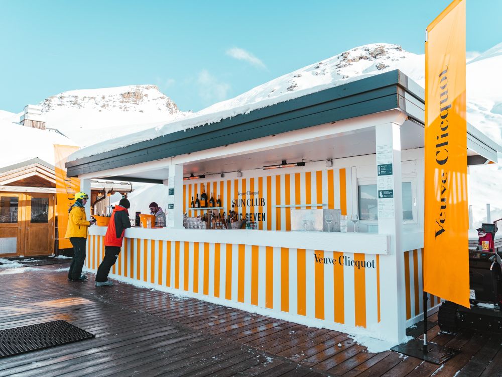 Bar extérieur Veuve Clicquot avec des rayures blanches et jaunes, deux personnes vêtues de vêtements de ski discutant avec un serveur, montagnes enneigées en arrière-plan sous un ciel bleu clair.