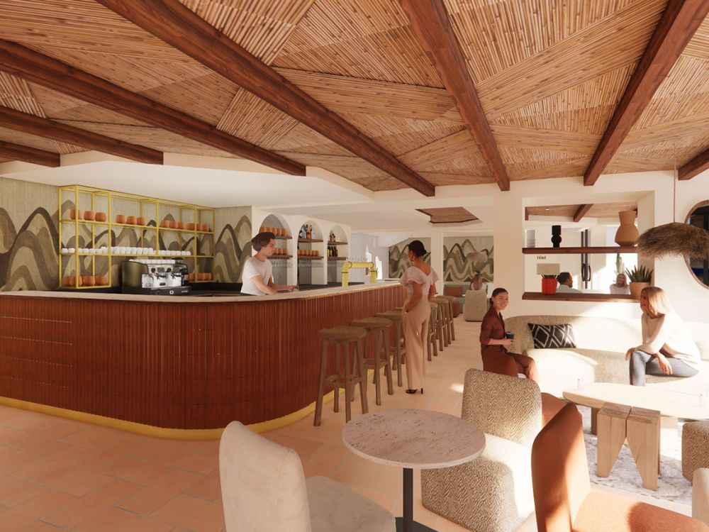 Intérieur lumineux d'un bar avec un grand comptoir en bois courbé, tabourets hauts en bois, plafond en bois tissé avec poutres apparentes, et mur avec motifs verts et jaunes. Plusieurs personnes sont présentes : une barista derrière le comptoir, une femme debout près des tabourets, et deux autres assises sur un canapé beige près d'une table basse. L'ambiance est chaleureuse avec des meubles en bois et des décorations modernes.