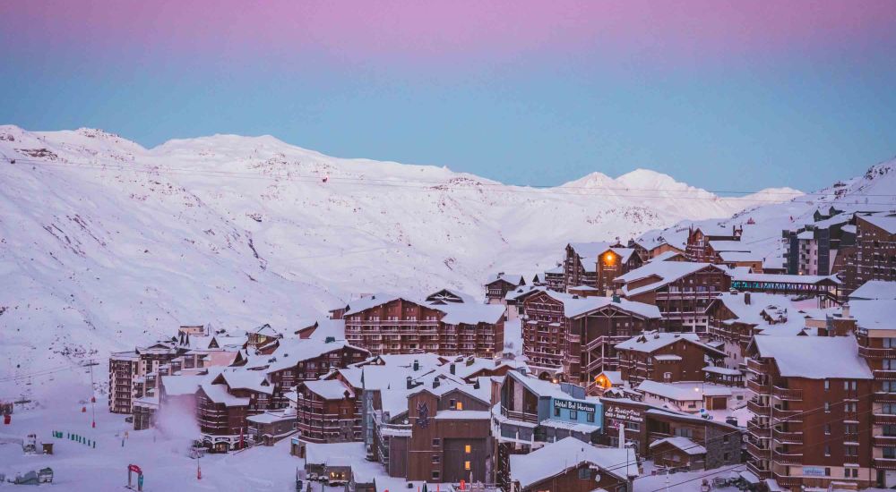 Vue panoramique d'une station de ski alpine au crépuscule avec des montagnes enneigées et des bâtiments en bois recouverts de neige sous un ciel rose et bleu.