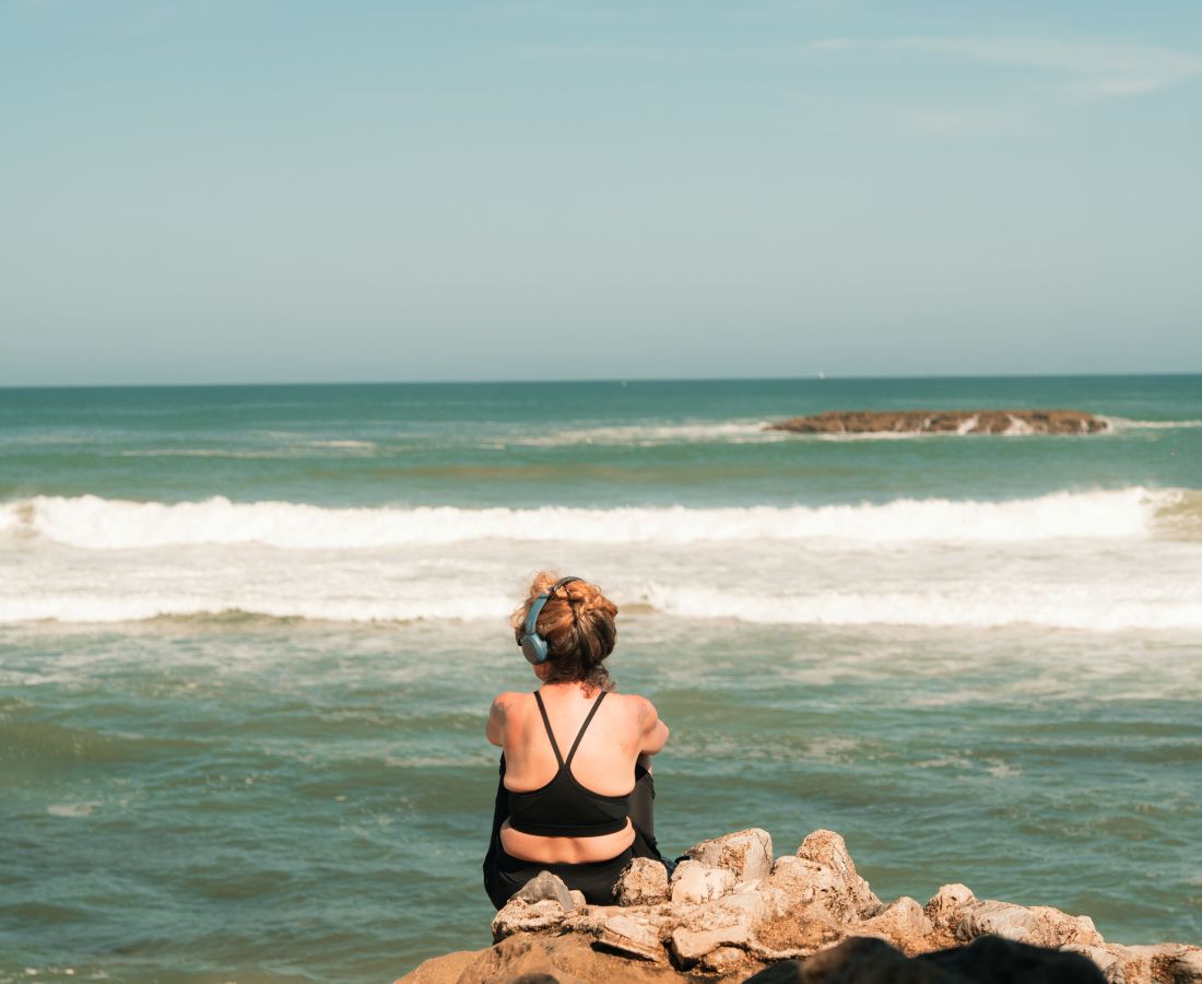 Une femme vue de dos assise sur des rochers, portant un haut de sport noir et des écouteurs, regardant la mer avec des vagues et un îlot rocheux à l'horizon sous un ciel bleu clair.