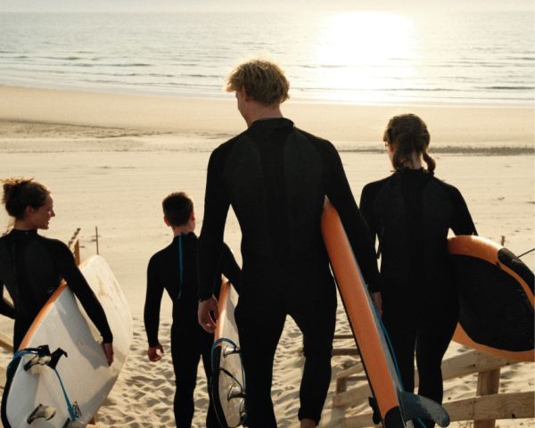 Quatre surfeurs portant des combinaisons noires portant des planches de surf orange et blanches marchant vers l'océan au coucher du soleil sur une plage de sable.