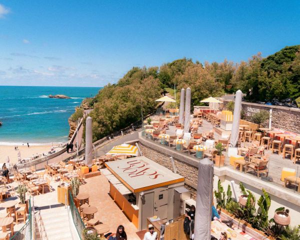 Terrasse extérieure d'un restaurant avec tables et chaises en bois surplombant une plage et l'océan sous un ciel bleu, entourée de végétation verte.
