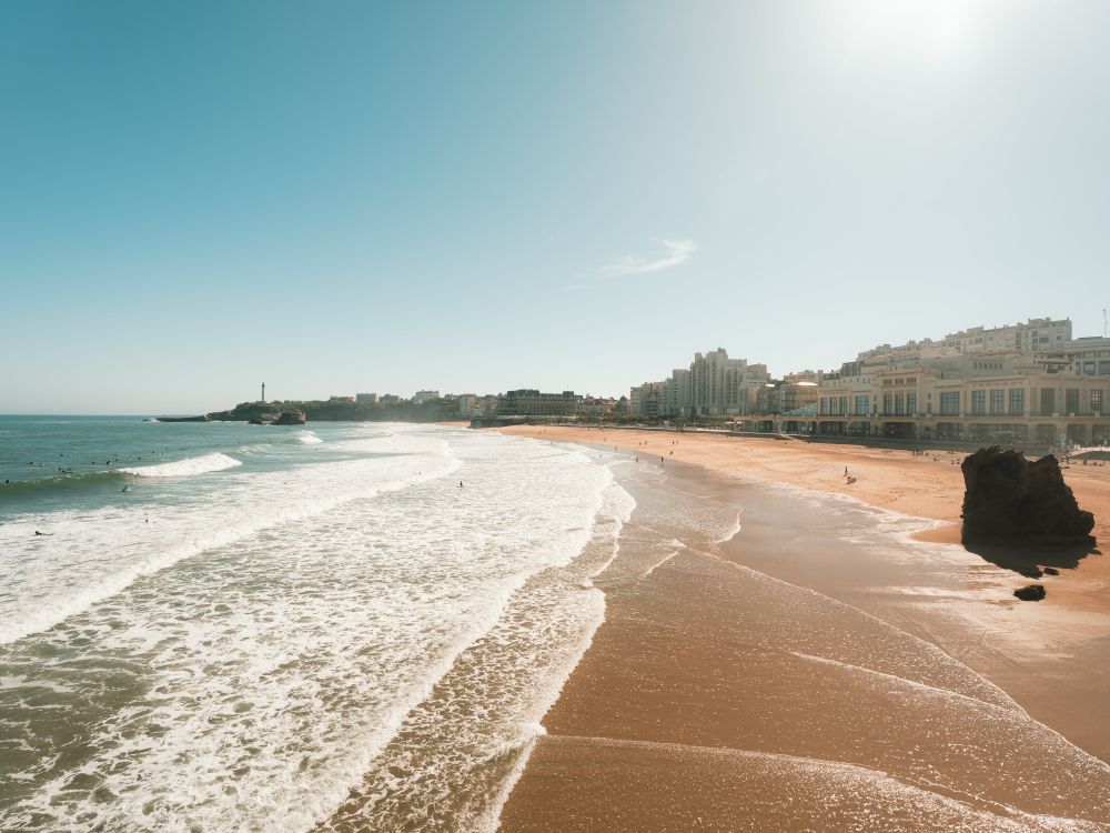 Plage de sable avec des vagues douces et des surfeurs dans l'eau, bâtiments en bord de mer et phare à l'arrière-plan sous un ciel clair.