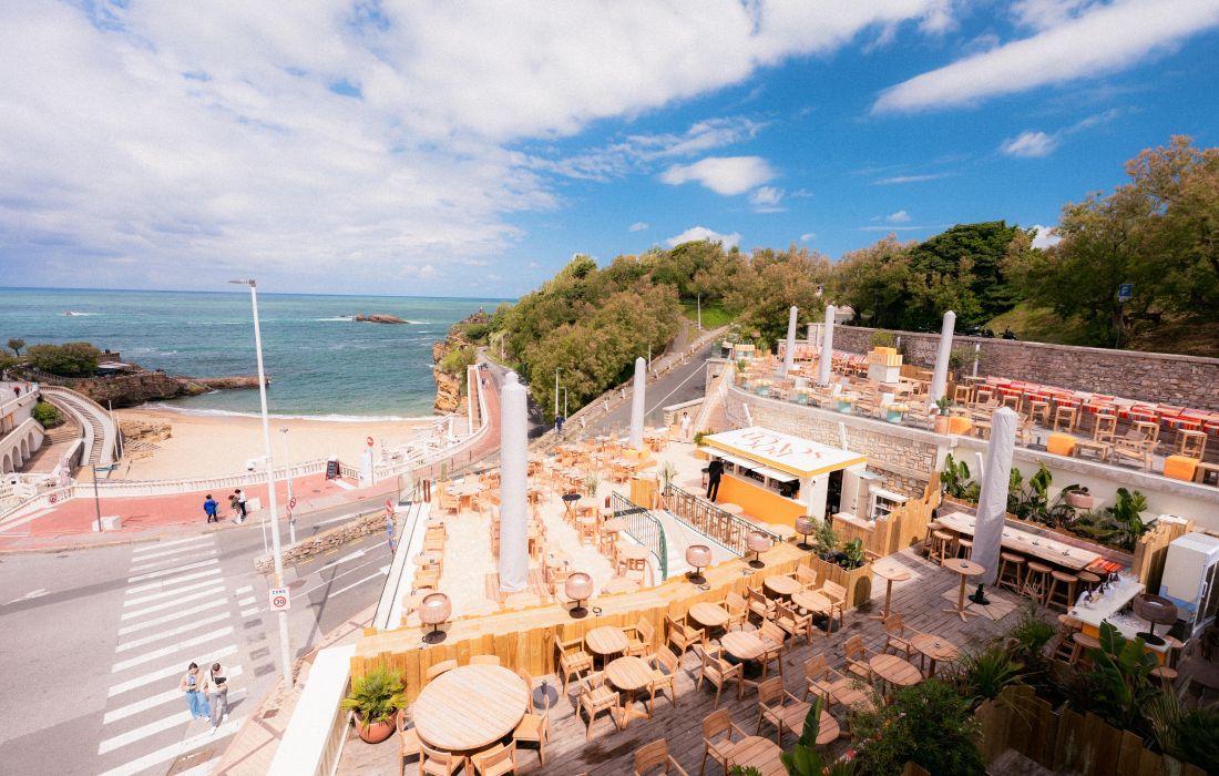Vue aérienne d'une terrasse de café avec de nombreuses tables et chaises en bois, située près d'une plage de sable et de l'océan sous un ciel partiellement nuageux.
