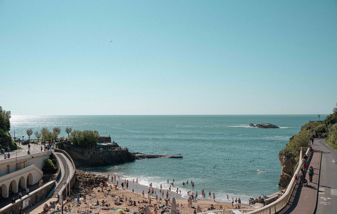 Plage animée avec des personnes bronzant et nageant, entourée de falaises, d'une promenade et de constructions en pierre sous un ciel bleu clair.