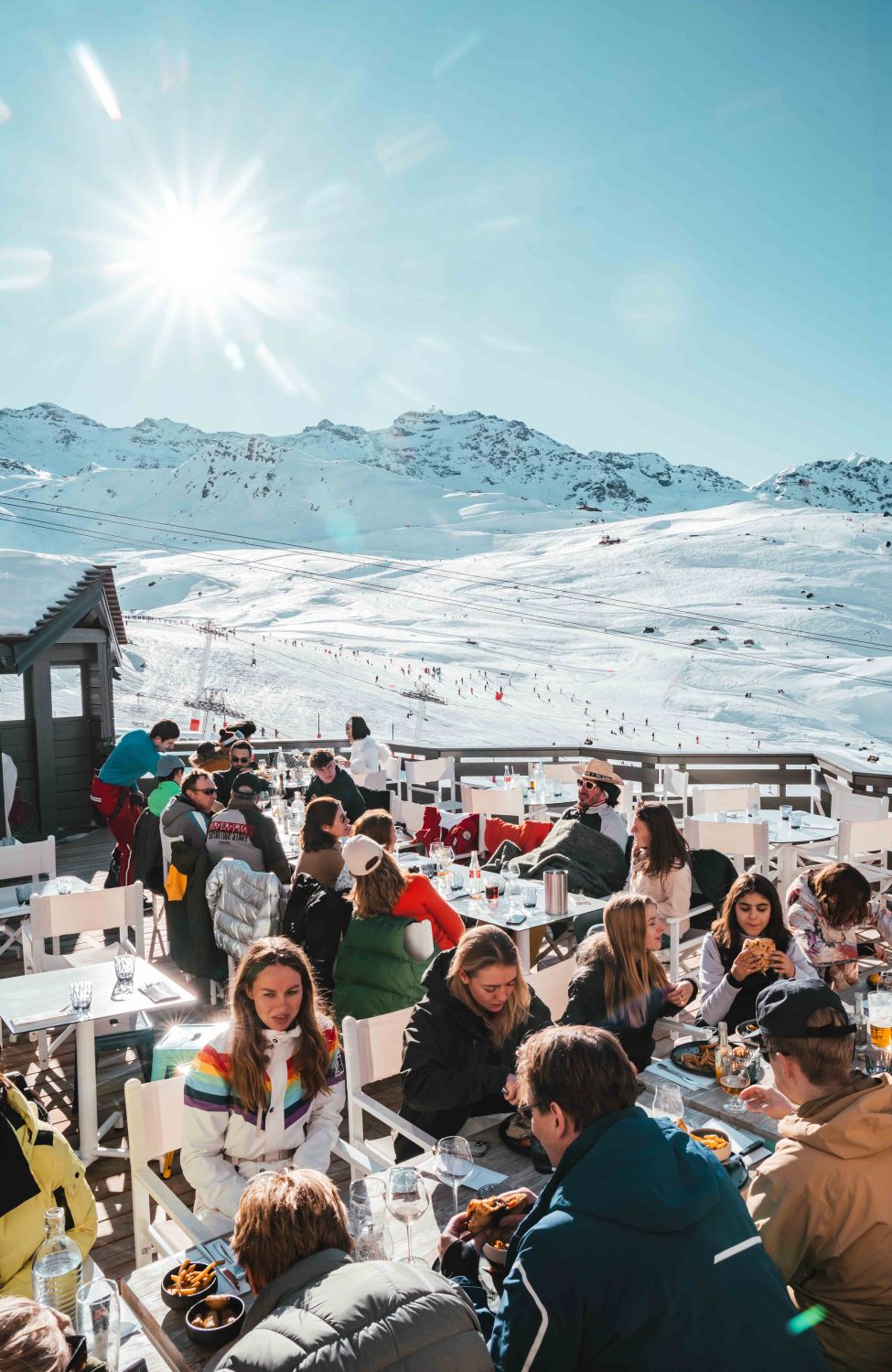 Groupe de personnes mangeant et discutant à une terrasse ensoleillée avec vue sur des montagnes enneigées et des skieurs en arrière-plan sous un ciel bleu clair.