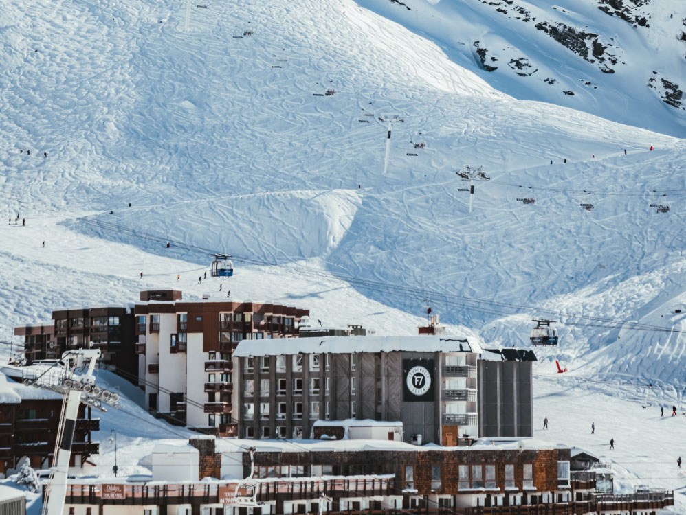 Station de ski avec des bâtiments modernes au premier plan et des pistes enneigées au fond où des skieurs et télésièges sont visibles sur la montagne.