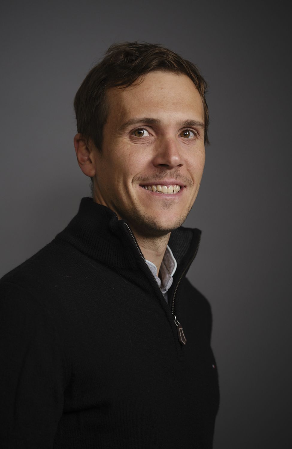 Portrait d'un homme souriant avec un pull noir à col zippé sur fond gris.