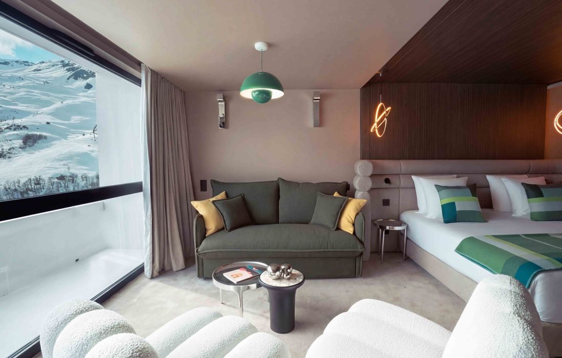 Chambre d'hôtel moderne avec un grand canapé vert orné de coussins jaunes et verts, un lit double avec des oreillers assortis, une lampe suspendue verte, une table basse et une fenêtre offrant une vue sur des montagnes enneigées.