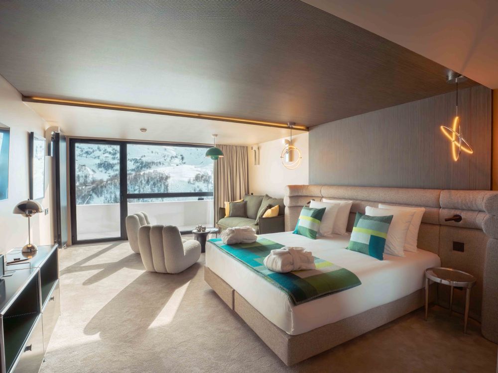 Chambre d'hôtel moderne avec un grand lit double doté de coussins aux tons verts et bleus, deux peignoirs pliés sur le lit, un fauteuil blanc moelleux, un canapé vert avec coussins, une grande baie vitrée offrant une vue sur un paysage enneigé de montagnes, des lampes suspendues modernes et une décoration épurée.