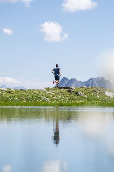Un homme court sur un sentier herbeux près d'un plan d'eau calme avec des montagnes en arrière-plan sous un ciel bleu avec quelques nuages.