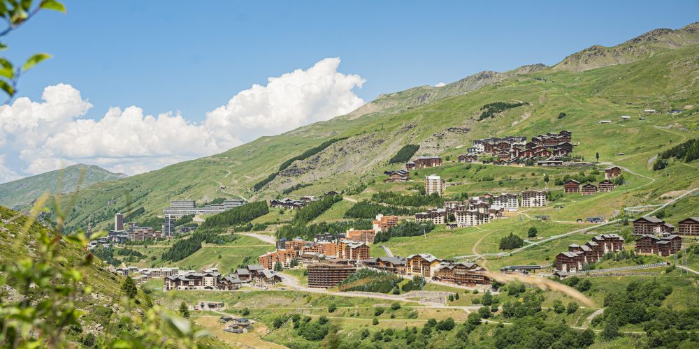 Vue panoramique d'une station de montagne en été avec de nombreux chalets et bâtiments répartis sur des pentes verdoyantes sous un ciel bleu avec quelques nuages blancs.