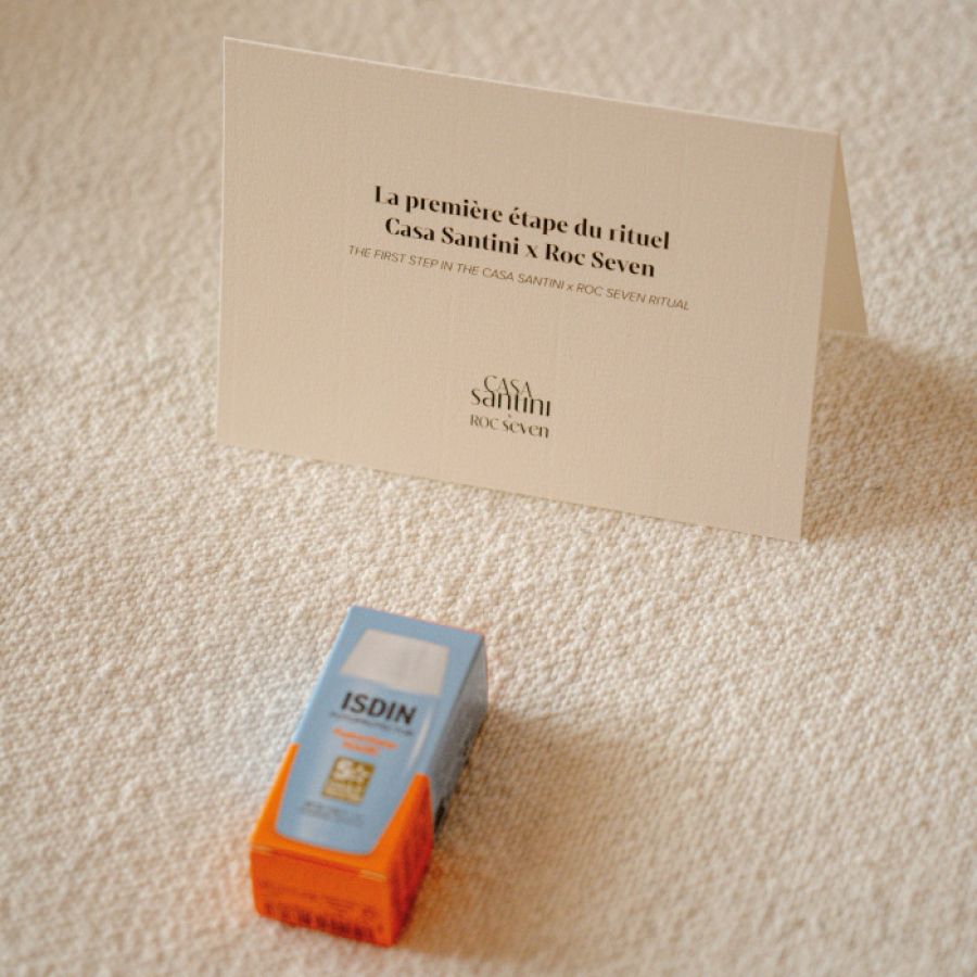Carte beige posée sur une surface texturée avec le texte "La première étape du rituel Casa Santini x Roc Seven" et une petite boîte de crème solaire ISDIN orange et bleue en avant-plan.