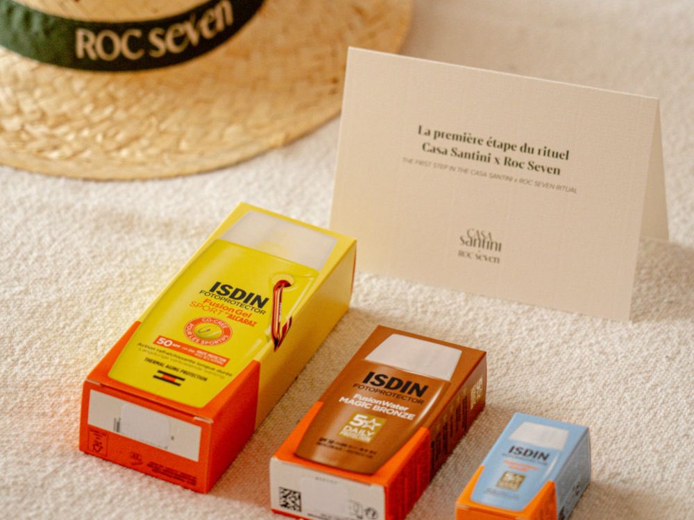 Trois boîtes de crème solaire ISDIN posées sur une surface texturée, avec un chapeau en paille portant l'inscription ROC Seven en arrière-plan et une carte pliée indiquant La première étape du rituel Casa Santini x Roc Seven.