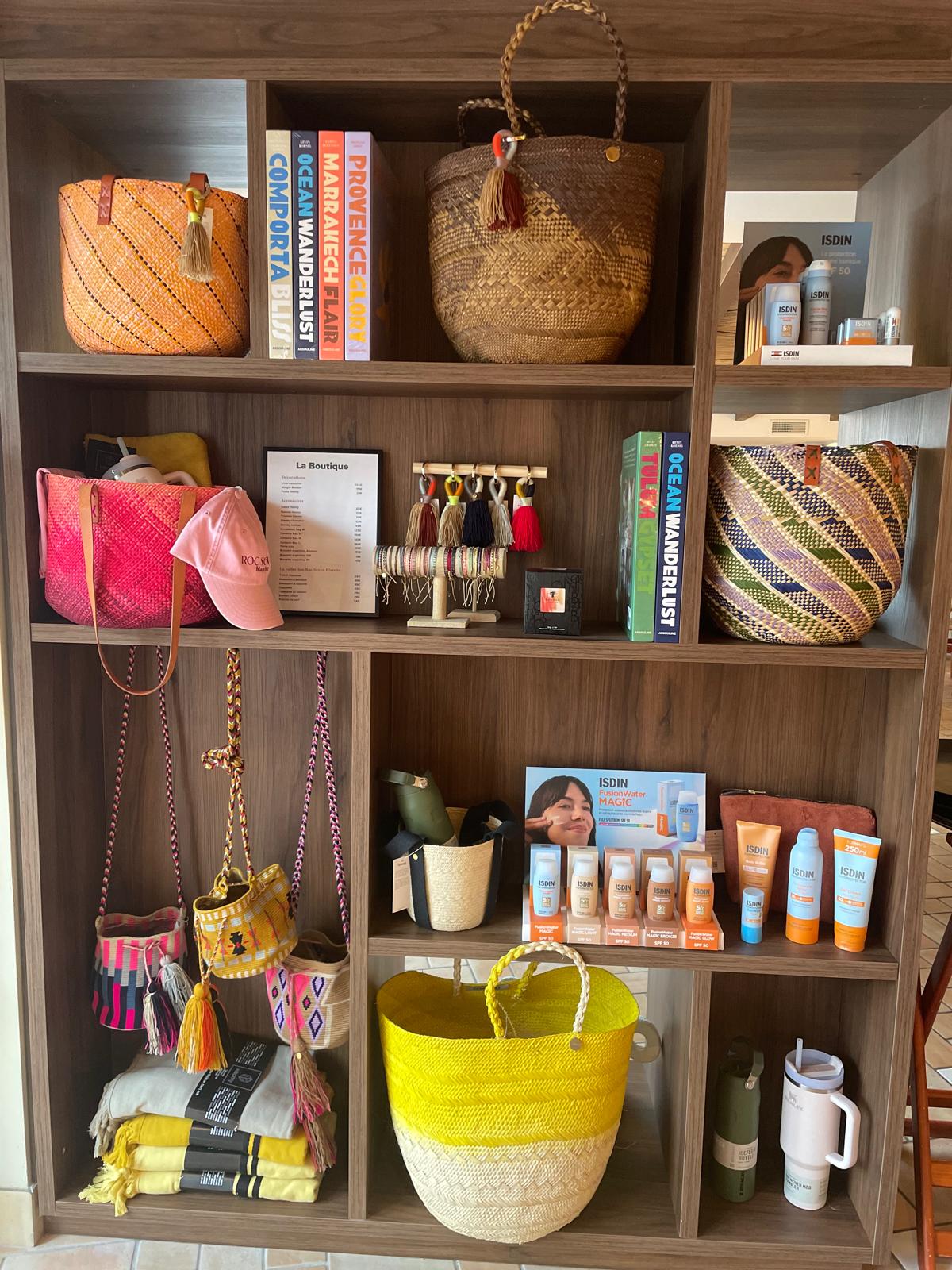 Étagère en bois avec plusieurs compartiments contenant des sacs tissés colorés, des livres de voyage, une casquette rose, des bracelets suspendus, des produits solaires ISDIN, des bouteilles et quelques foulards pliés.