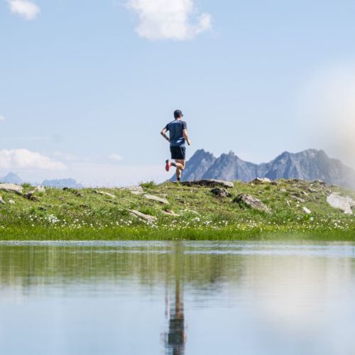 Un coureur en tenue de sport bleue court sur un sentier herbeux près d'un lac en montagne avec des sommets rocheux en arrière-plan sous un ciel bleu partiellement nuageux.