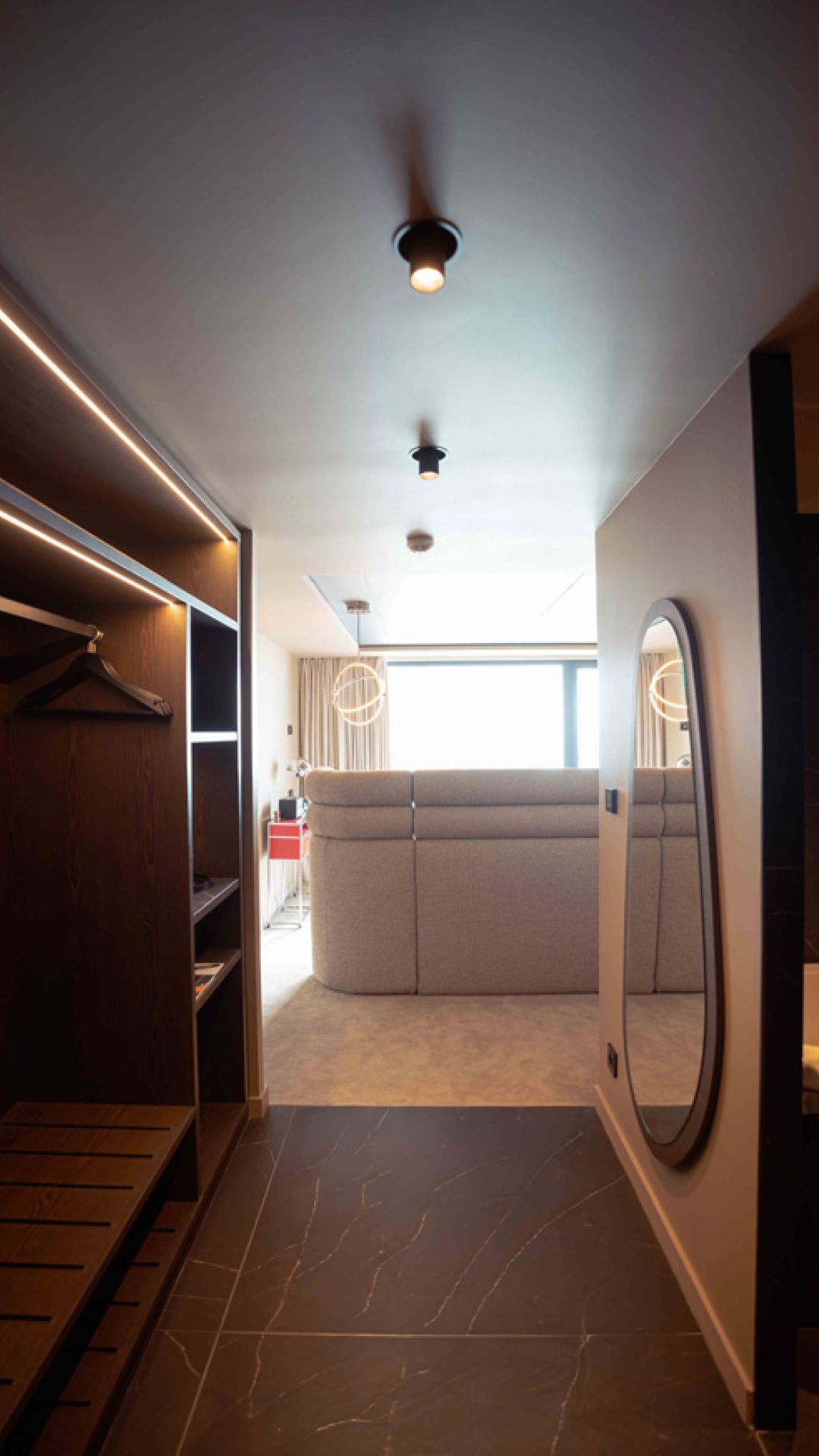 Entrée d'une chambre d'hôtel moderne avec un sol en carrelage sombre, un placard en bois avec des cintres à gauche, un grand miroir ovale fixé au mur à droite, et un salon lumineux avec un canapé gris devant une grande fenêtre au fond.
