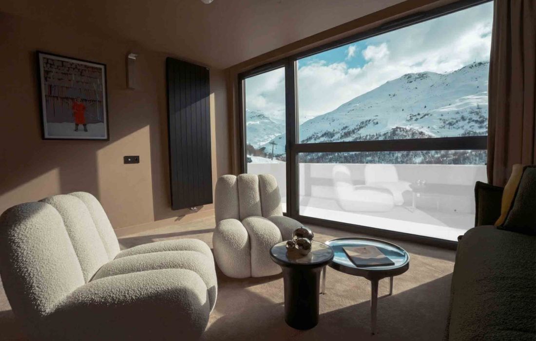 Salon moderne avec deux fauteuils en tissu texturé crème, petite table noire avec décorations métalliques, grande fenêtre offrant une vue sur des montagnes enneigées sous un ciel partiellement nuageux.