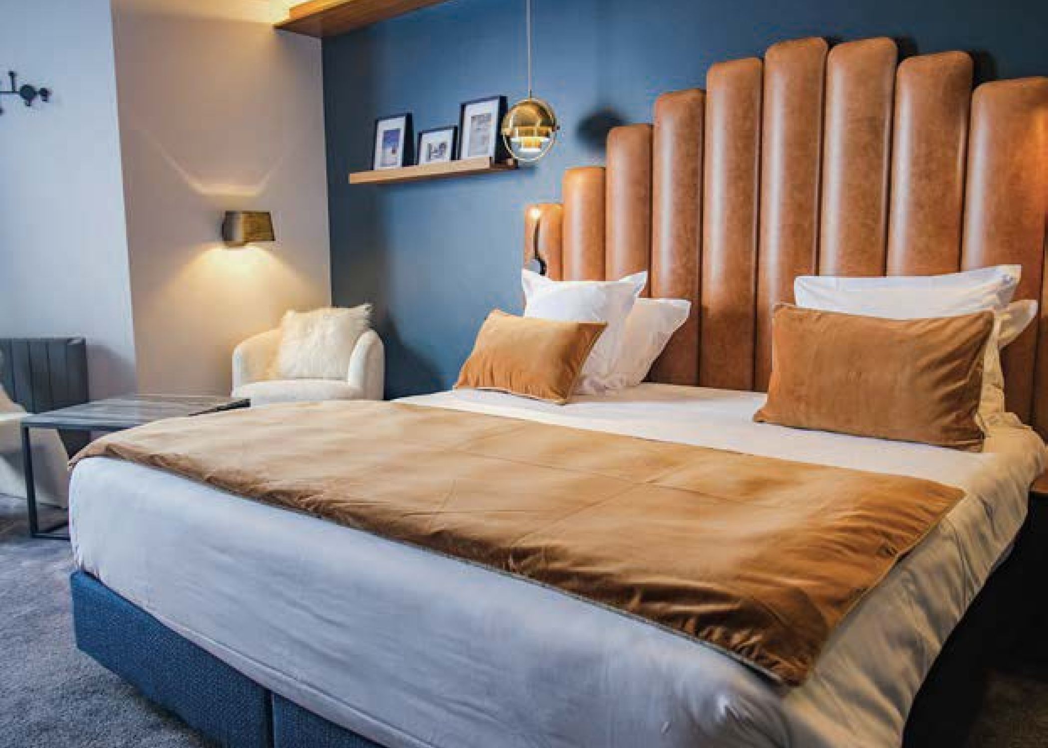 Chambre avec un grand lit double couvert d'une couette beige, des oreillers blancs et deux coussins marron, une tête de lit en cuir marron côtelée, un mur bleu foncé avec une étagère en bois portant trois cadres photo, une suspension dorée, un fauteuil blanc avec un coussin et une lampe murale allumée.