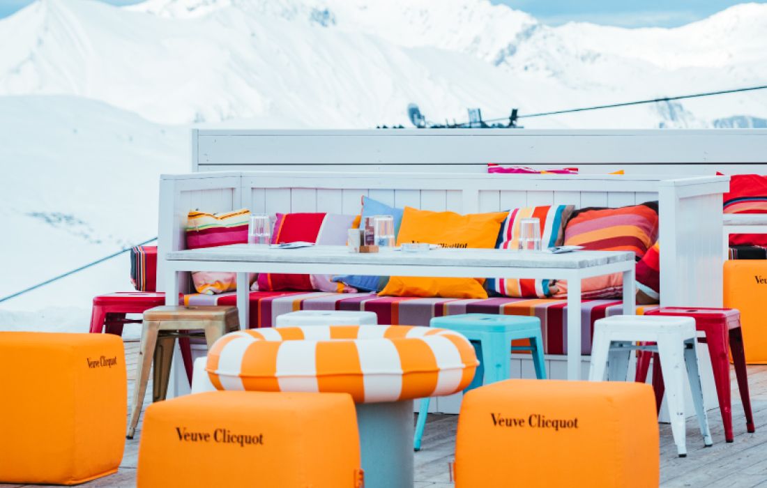 Espace terrasse extérieur avec sièges et coussins colorés autour d'une table, avec des poufs orange marqués Veuve Clicquot, et un paysage de montagnes enneigées en arrière-plan.