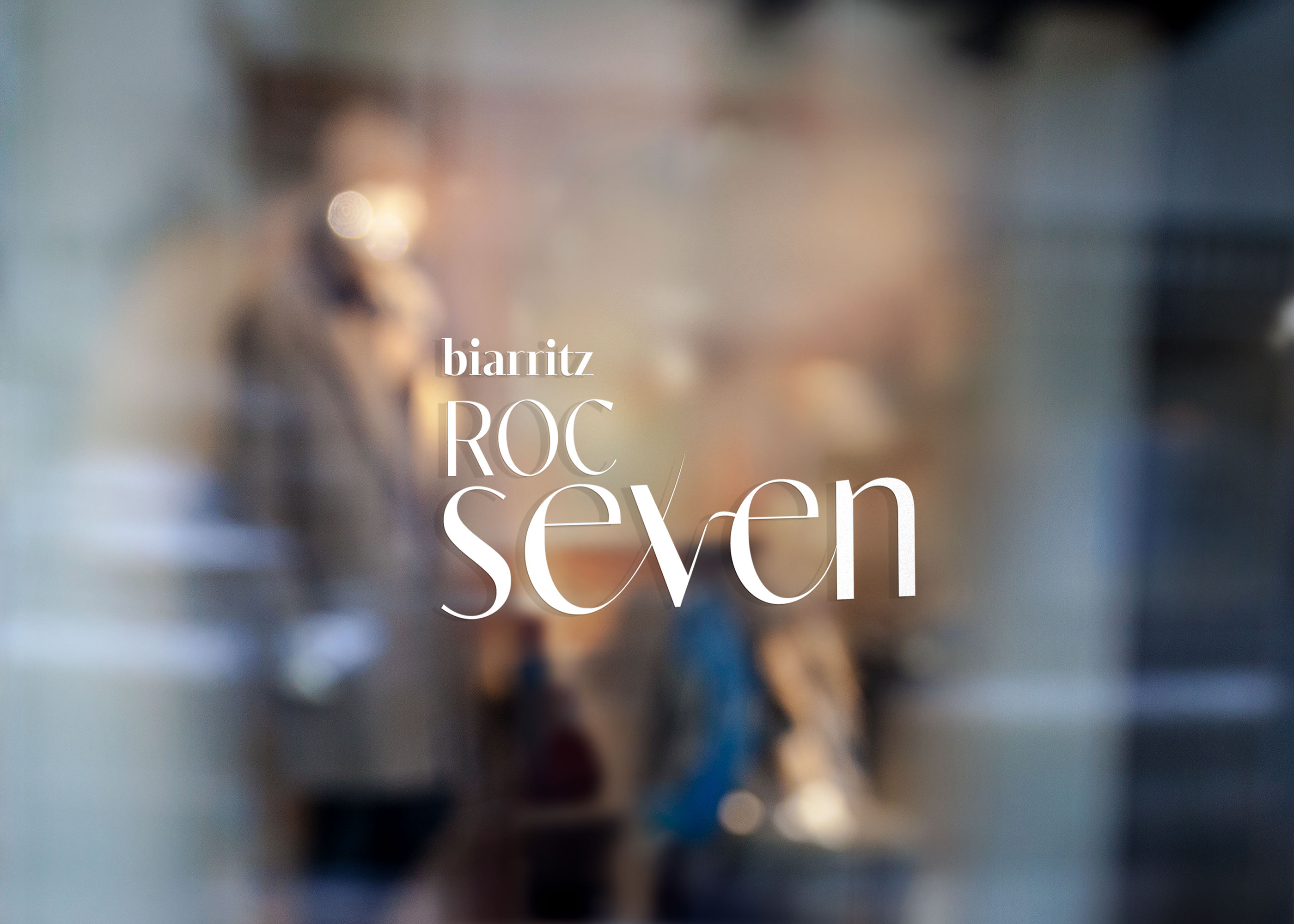 Vitrophanie sur une vitrine avec le texte "biarritz ROC seven" en blanc sur un fond flou montrant une silhouette et des objets à l'intérieur.