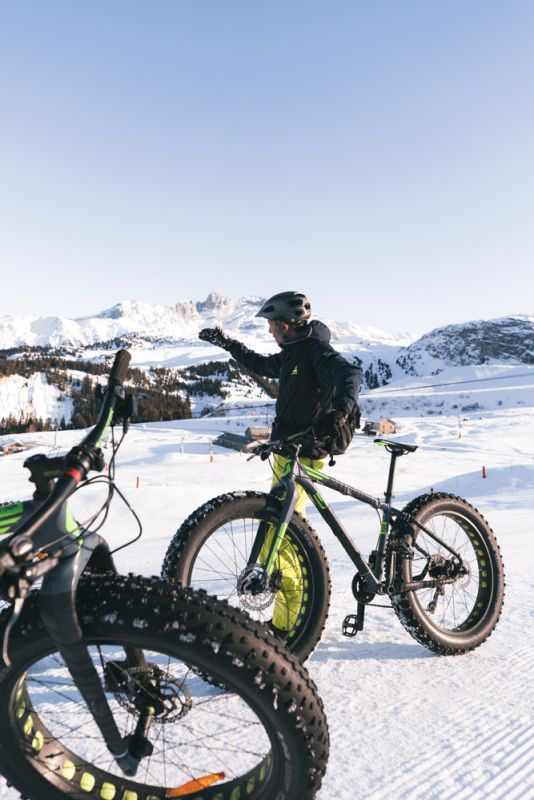 Personne vêtue d'un casque et d'une tenue de sport sur un vélo à gros pneus sur un sentier enneigé avec des montagnes enneigées en arrière-plan sous un ciel bleu clair.
