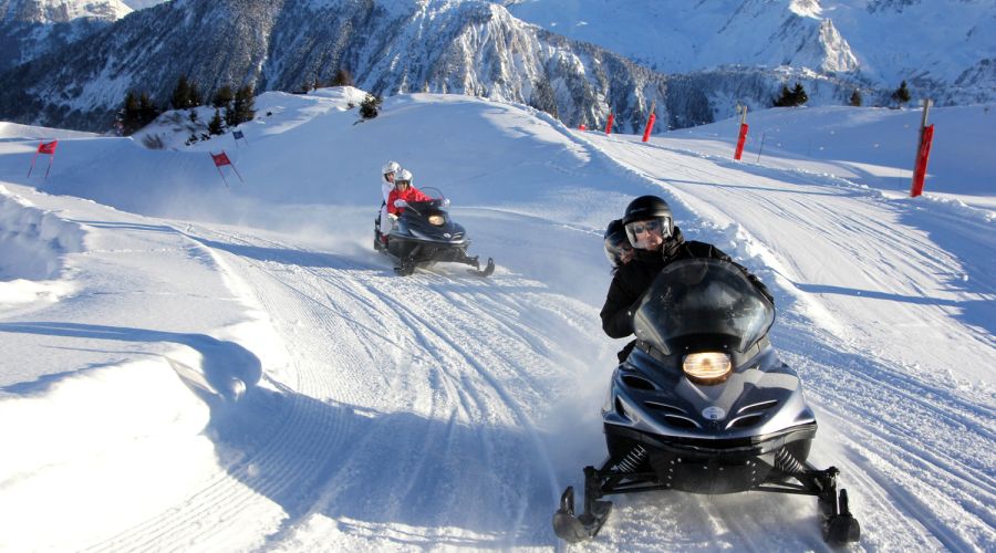 Deux motoneiges avec des conducteurs et passagers portant des casques roulent sur une piste enneigée dans un paysage montagneux ensoleillé.