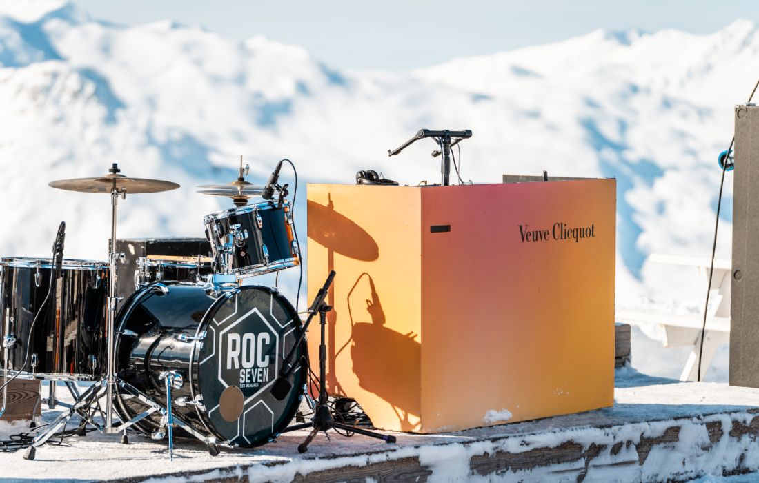 Batterie de musique noire avec logo ROC SEVEN posée sur une terrasse enneigée, à côté d'un pupitre orange Veuve Clicquot, avec des montagnes enneigées en arrière-plan.