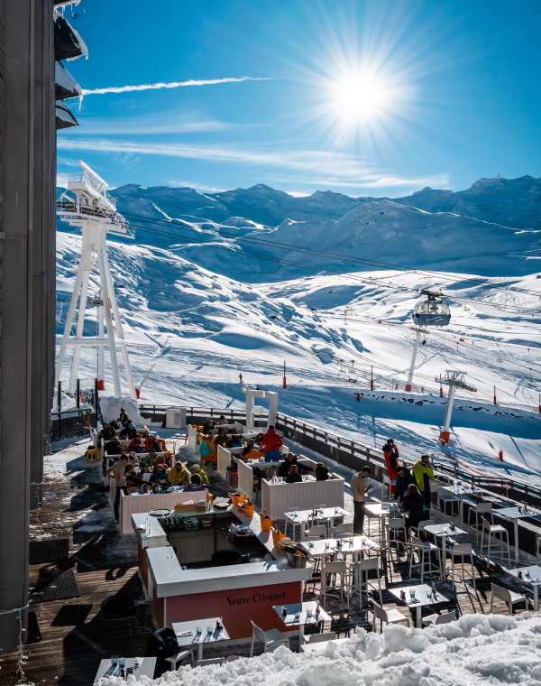 Terrasse ensoleillée d'un bar en montagne avec des clients assis, entourée de neige, avec un téléphérique et des montagnes enneigées en arrière-plan sous un ciel bleu clair.
