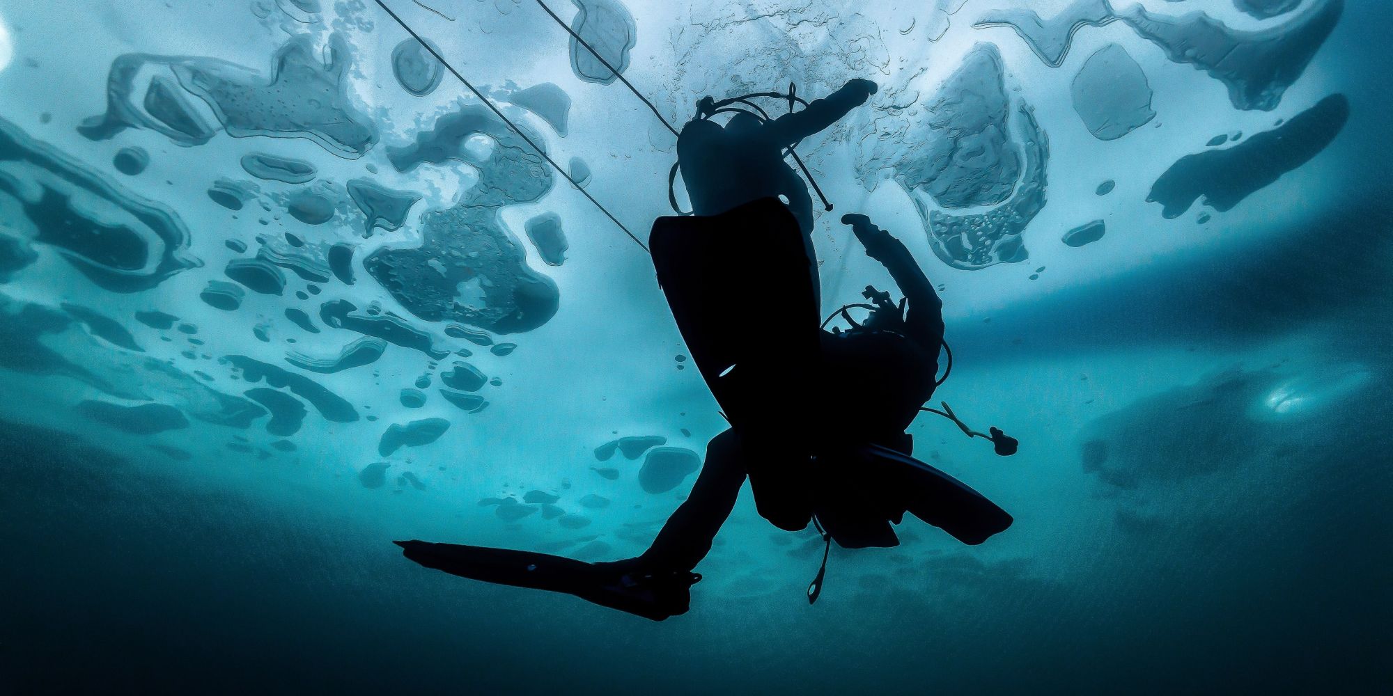 Un plongeur silhouette vu de dessous nage sous une surface gelée avec des formations de glace translucides au-dessus de lui.