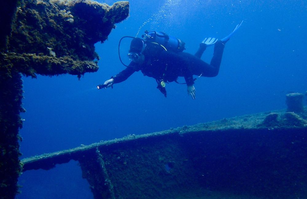 Plongeur en combinaison noire avec palmes bleues explorant l'épave d'un navire recouvert d'algues et de coraux sous l'eau.