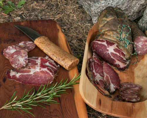Assiette en bois avec des tranches de charcuterie corse, un couteau à manche en bois, et un brin de romarin posé à côté sur une planche en bois.