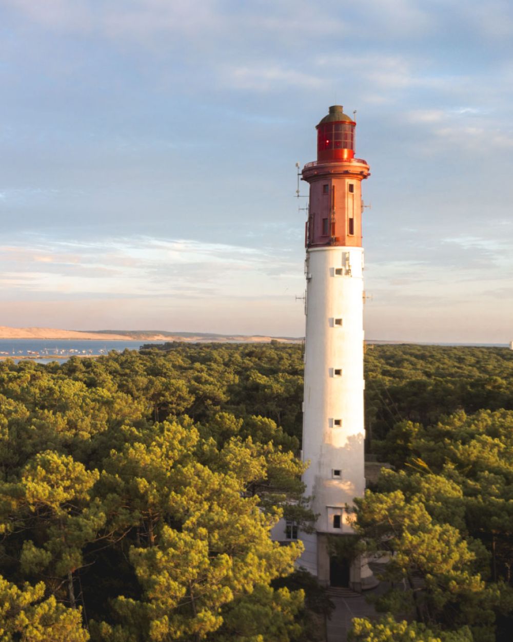 Phare blanc et rouge surplombant une forêt dense de pins avec un plan d'eau et des dunes à l'arrière-plan sous un ciel partiellement nuageux au coucher du soleil.