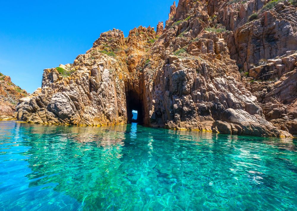 Eaux turquoise claires menant à une arche rocheuse naturelle dans des falaises escarpées sous un ciel bleu clair.
