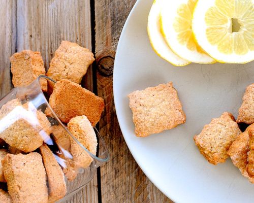 Biscuits carrés croustillants sur une assiette blanche à côté de tranches de citron et un verre renversé avec plus de biscuits sur une table en bois.