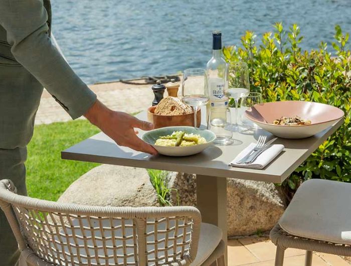 Une table carrée en extérieur près de l'eau avec deux chaises en osier, une bouteille de vin blanc, deux verres à vin, un plateau avec du pain, et deux plats servis, avec une personne posant un bol contenant un plat sur la table sous un ciel clair.