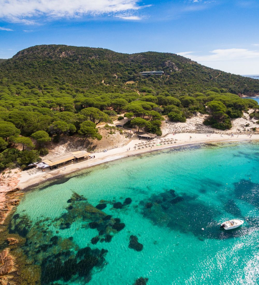 Vue aérienne d'une plage au sable clair bordée de parasols, une petite embarcation blanche dans une eau turquoise transparente avec des rochers visibles dessous, et une forêt dense de pins verts montant vers une colline boisée sous un ciel bleu partiellement nuageux.