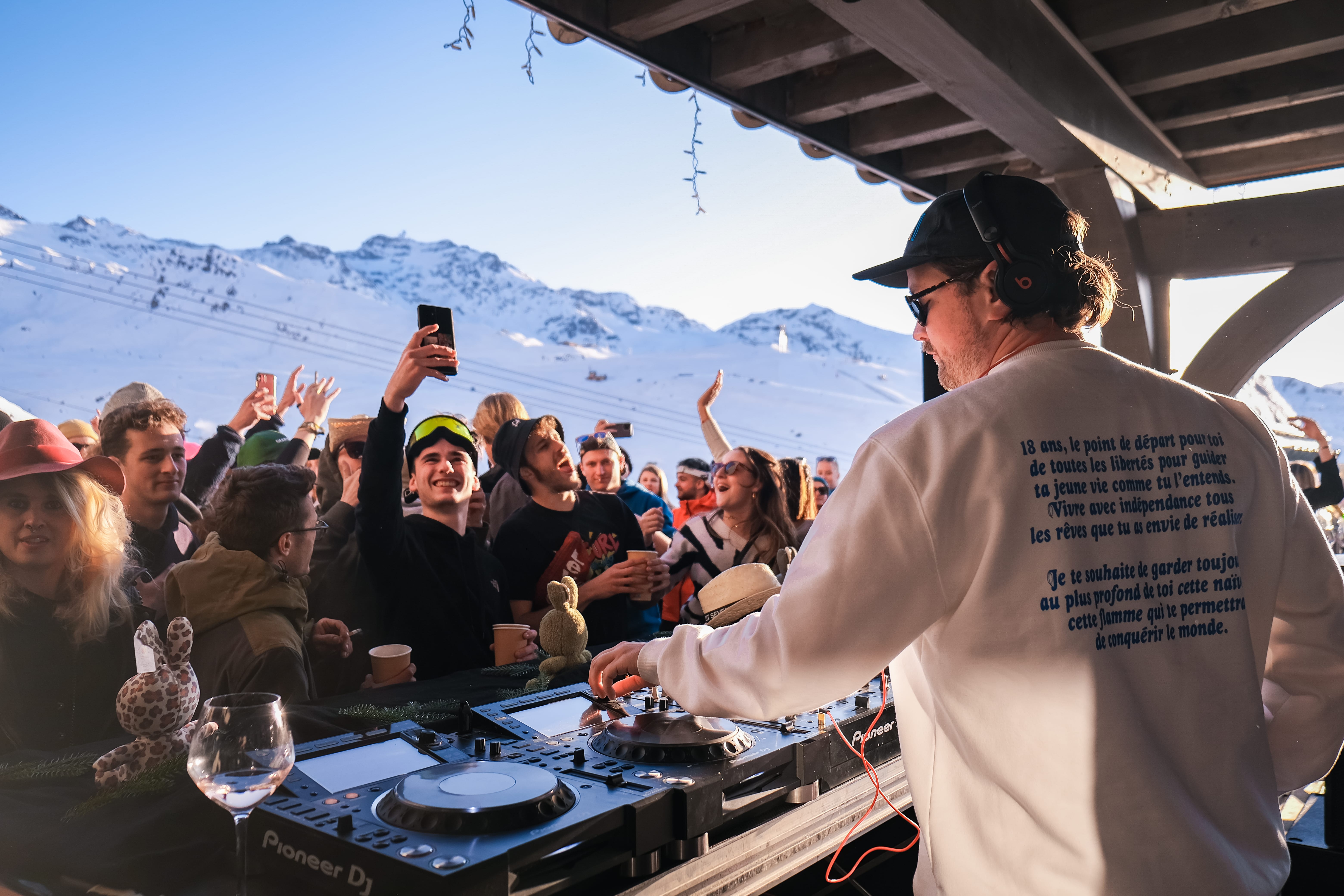 DJ en sweat-shirt blanc avec casque et lunettes de soleil anime une fête en plein air en montagne sous un abri en bois, avec un public joyeux, certains prenant des selfies et tenant des boissons, montagnes enneigées en arrière-plan sous un ciel bleu.