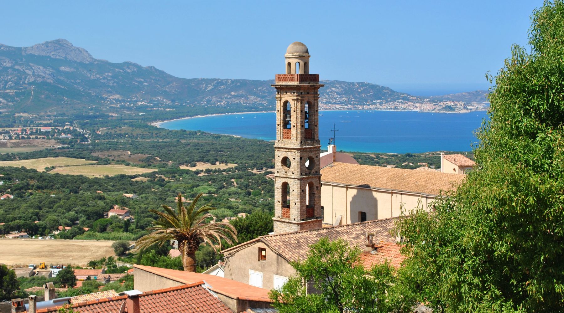 Tour de clocher en pierre avec des cloches, entourée de bâtiments aux toits de tuiles et de végétation, avec une vue panoramique sur une baie bleue, une étendue verte et des montagnes à l'arrière-plan sous un ciel bleu clair.