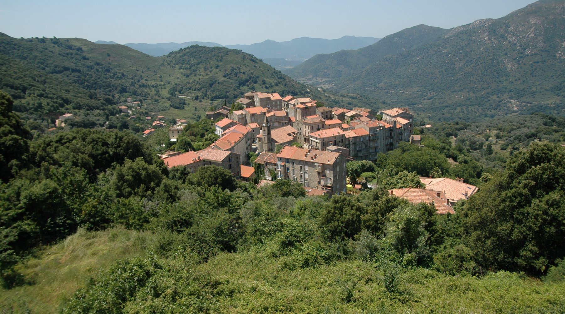 Petit village avec des maisons en pierre et toits en tuiles rouges entouré de collines verdoyantes et de montagnes sous un ciel bleu clair.