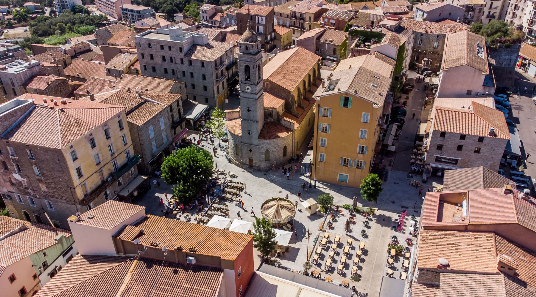 Place animée entourée de bâtiments aux toits en tuiles rouges, avec une église au clocher imposant au centre et des terrasses de cafés avec du mobilier en plein air.