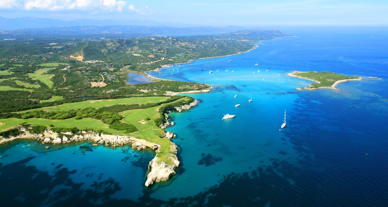 Vue aérienne d'une côte méditerranéenne avec des eaux turquoise, des petits bateaux à voile et à moteur, une péninsule verdoyante avec des rochers blancs, et des collines boisées en arrière-plan sous un ciel ensoleillé partiellement couvert de nuages.