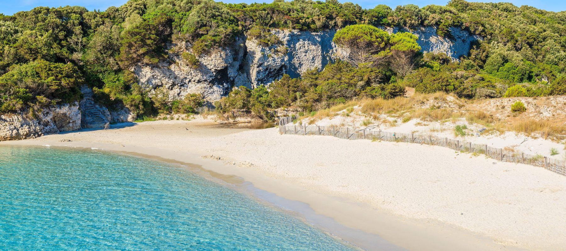 Plage au sable blanc bordée d'une eau turquoise claire avec une falaise rocheuse recouverte de végétation dense en arrière-plan sous un ciel bleu clair.