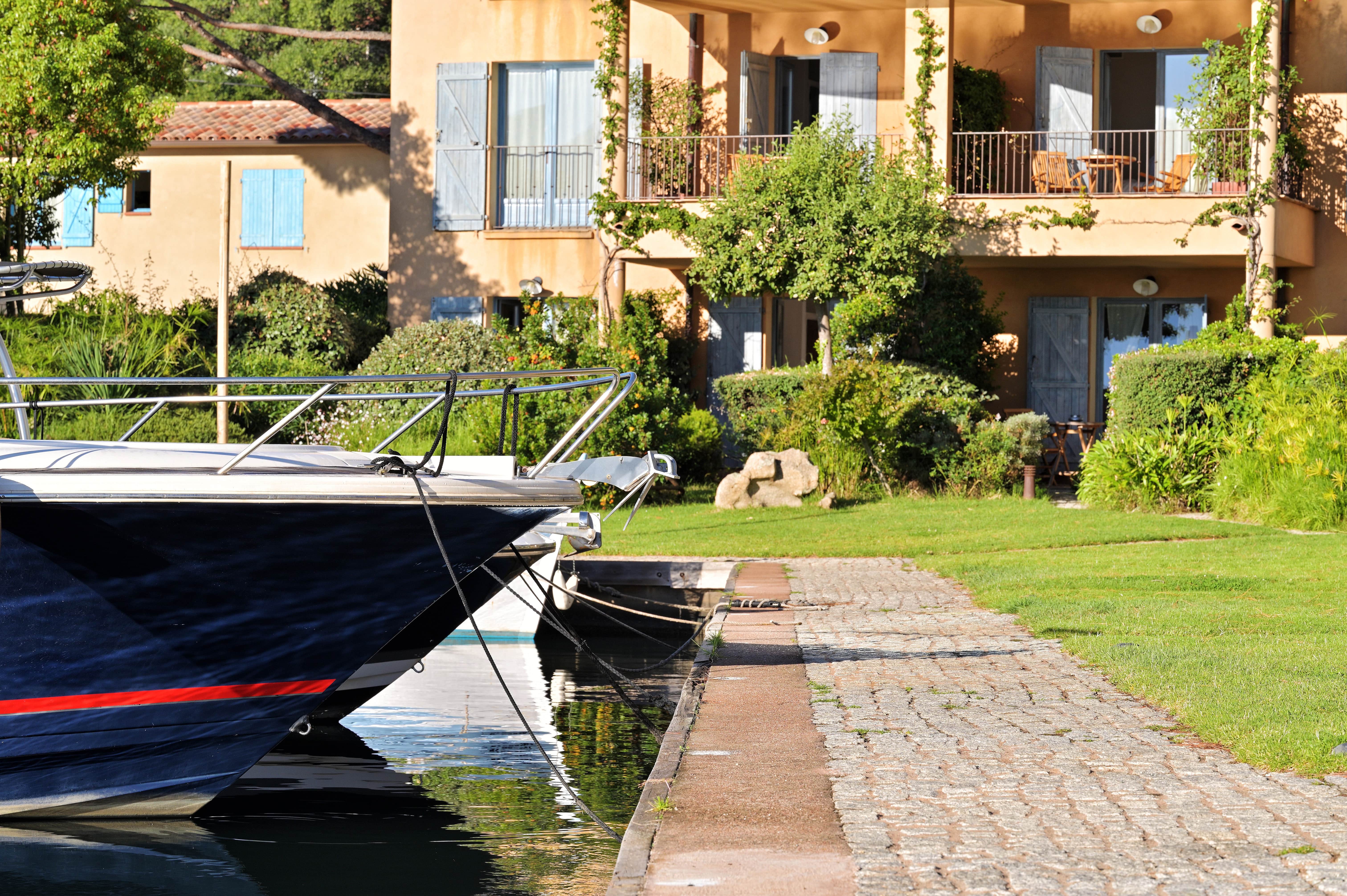 Bord d'eau avec un bateau amarré devant une maison beige aux volets bleus, entourée de buissons et d'arbres, avec une terrasse et une table en bois sur le balcon.