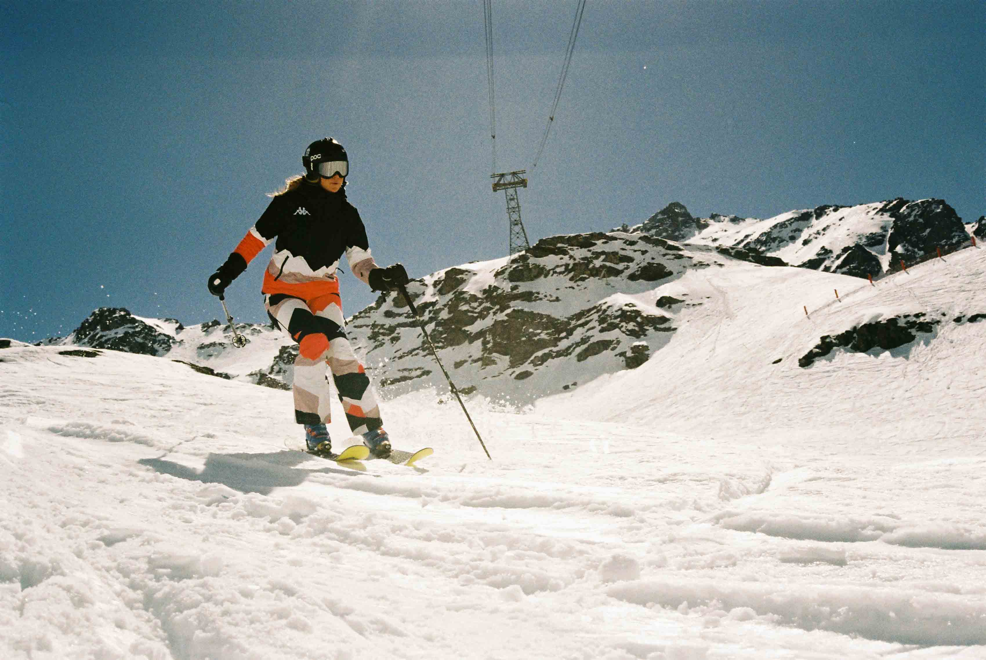Une skieuse descend une piste enneigée entourée de montagnes rocheuses sous un ciel bleu clair, portant une combinaison multicolore noire, orange et blanche avec un casque et des lunettes de ski.